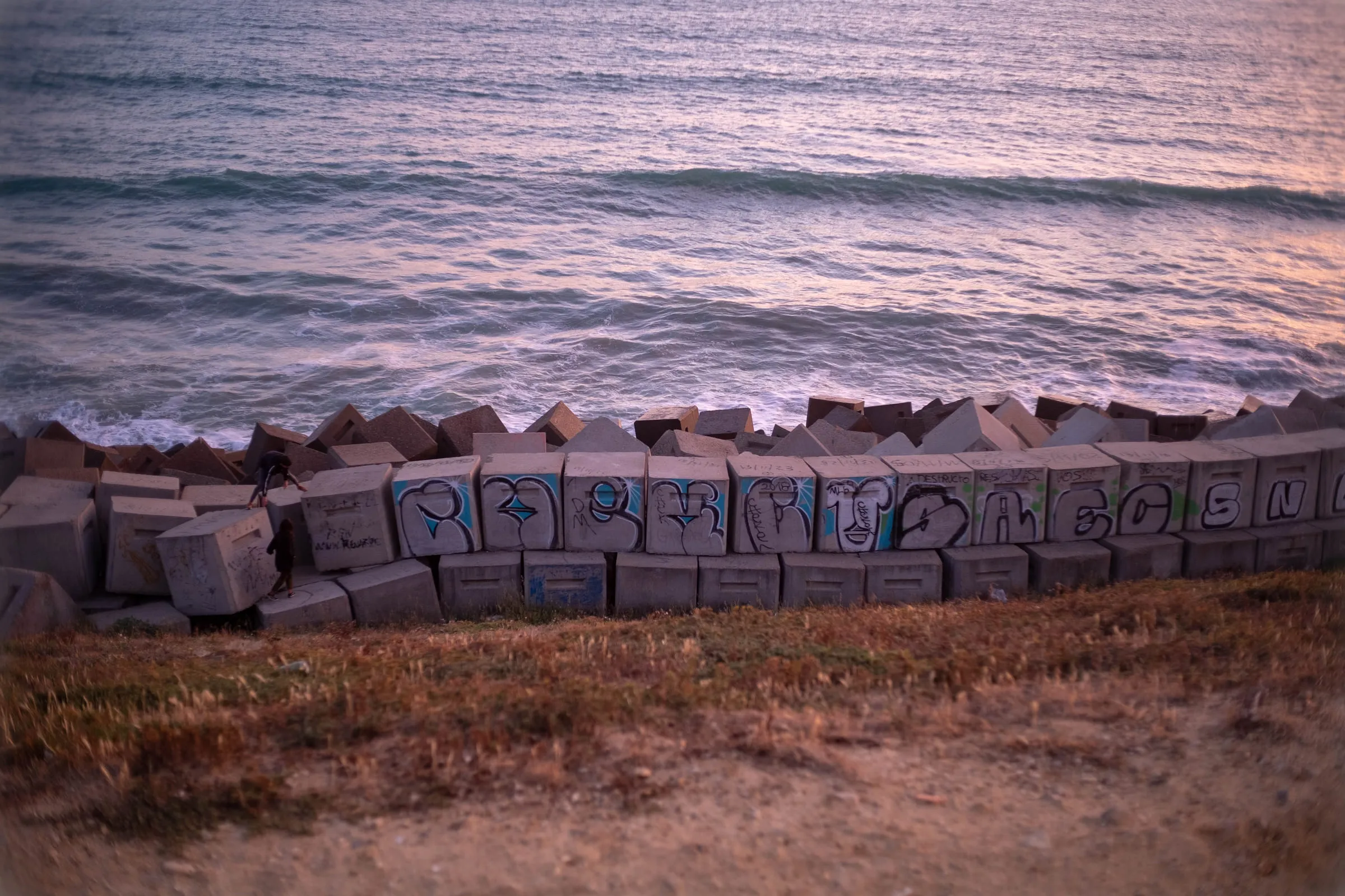 Graffitied concrete sea defence blocks piled along the shoreline, waves breaking beyond
