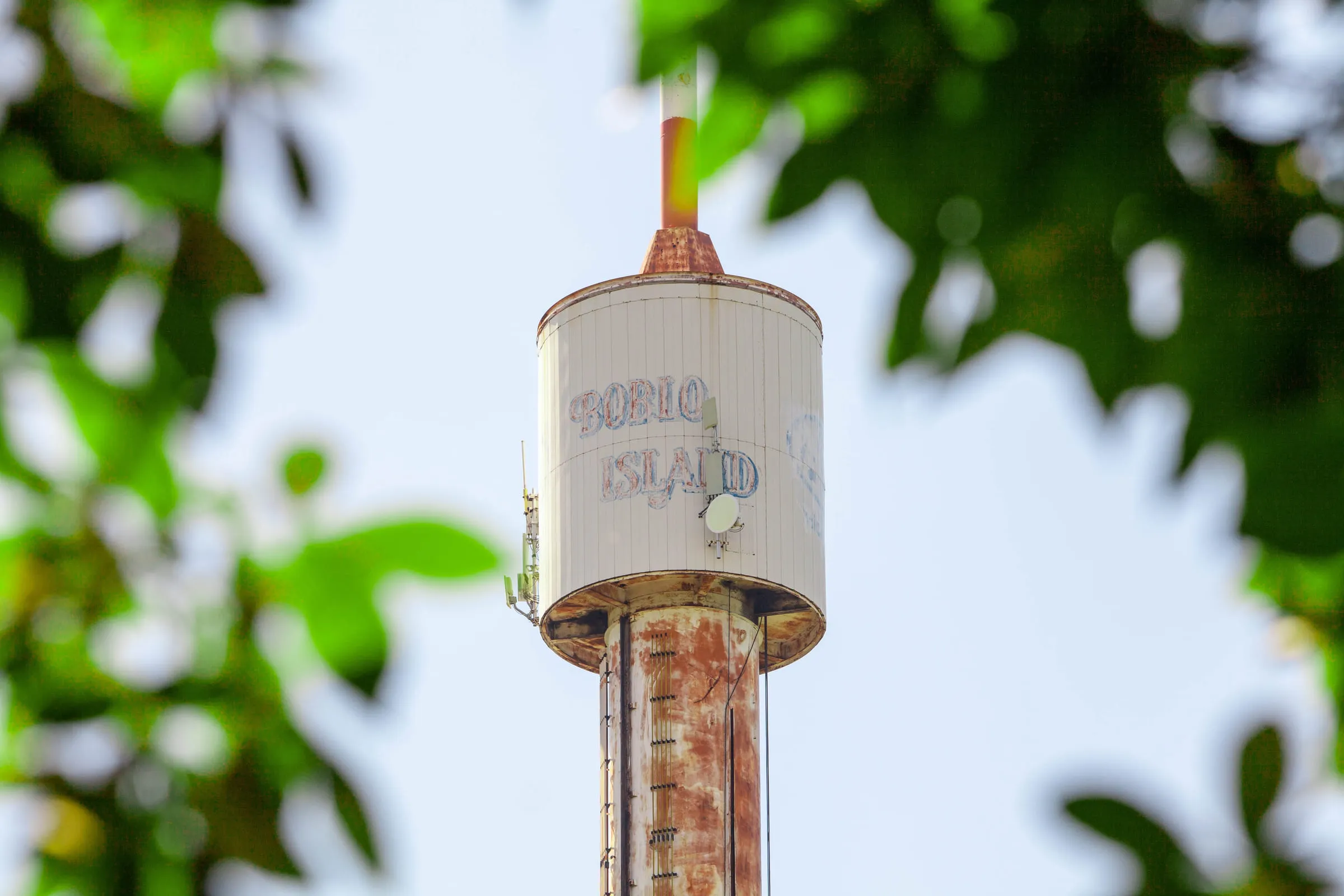 The top of the sky tower, its BOBLO ISLAND lettering weathered, framed by out-of-focus leaves