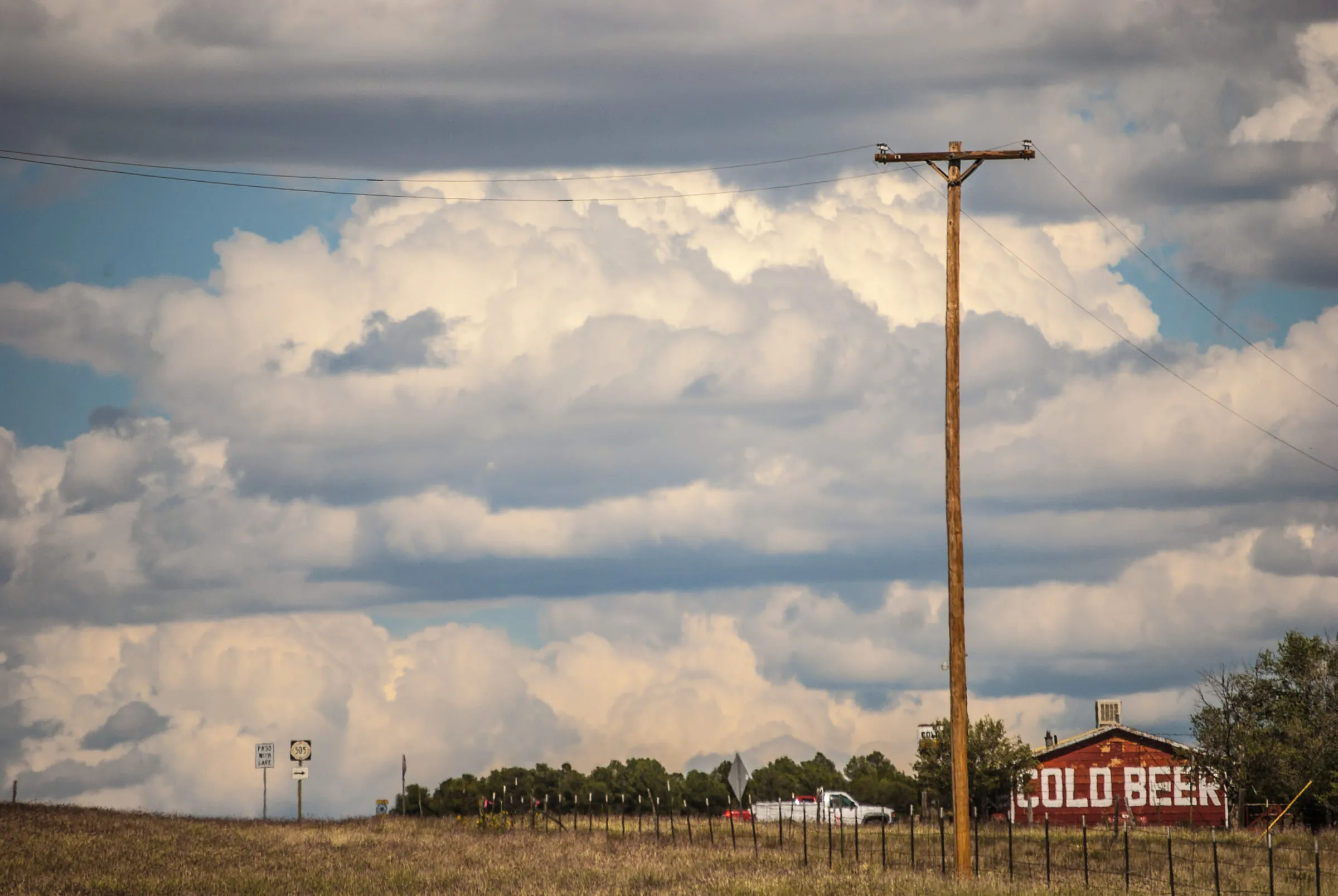 A leaning power pole in tall grass, the COLD BEER barn small on the horizon under piled clouds