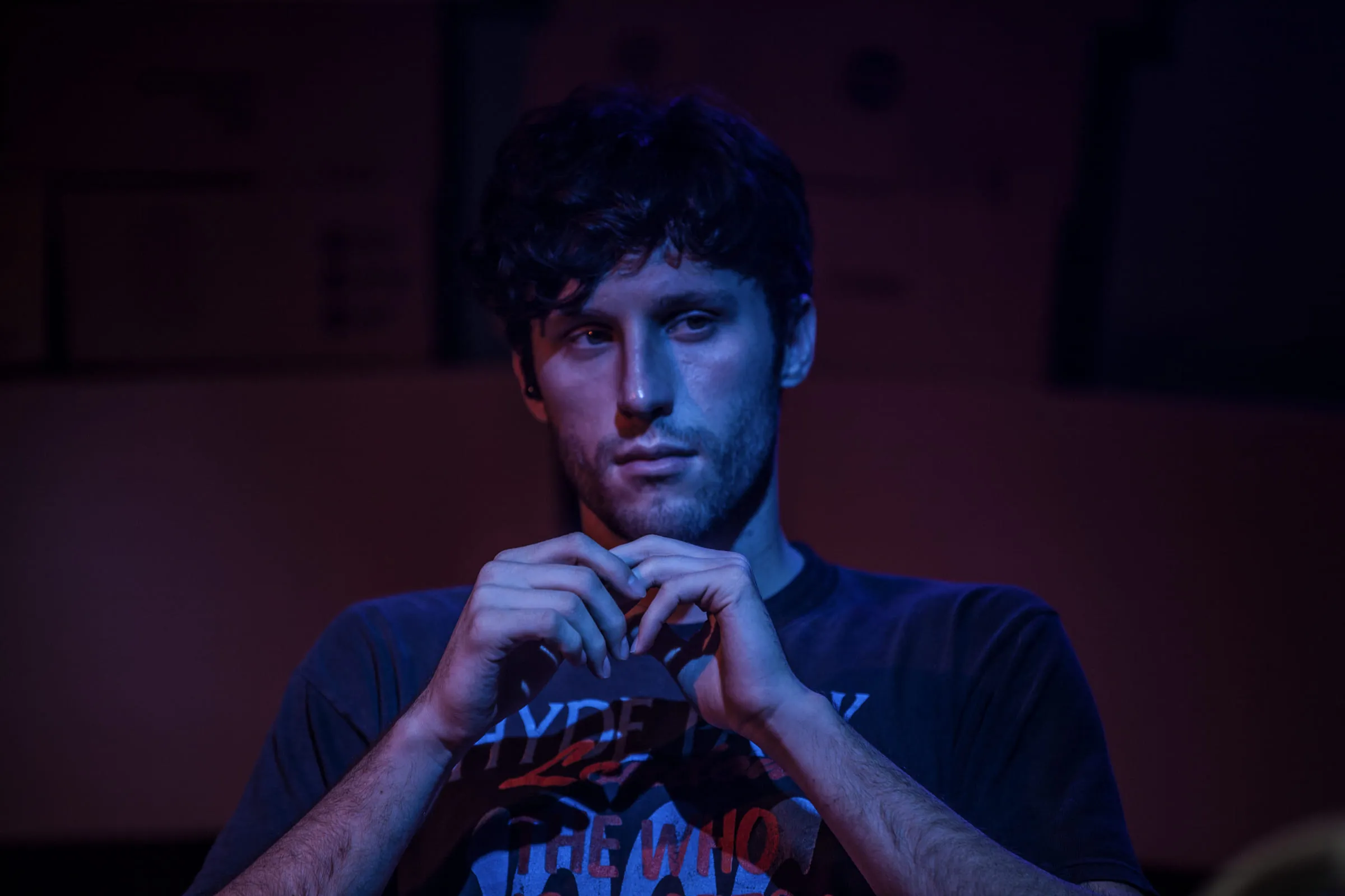 Collin seated under blue and magenta stage light, hands together in front of the face