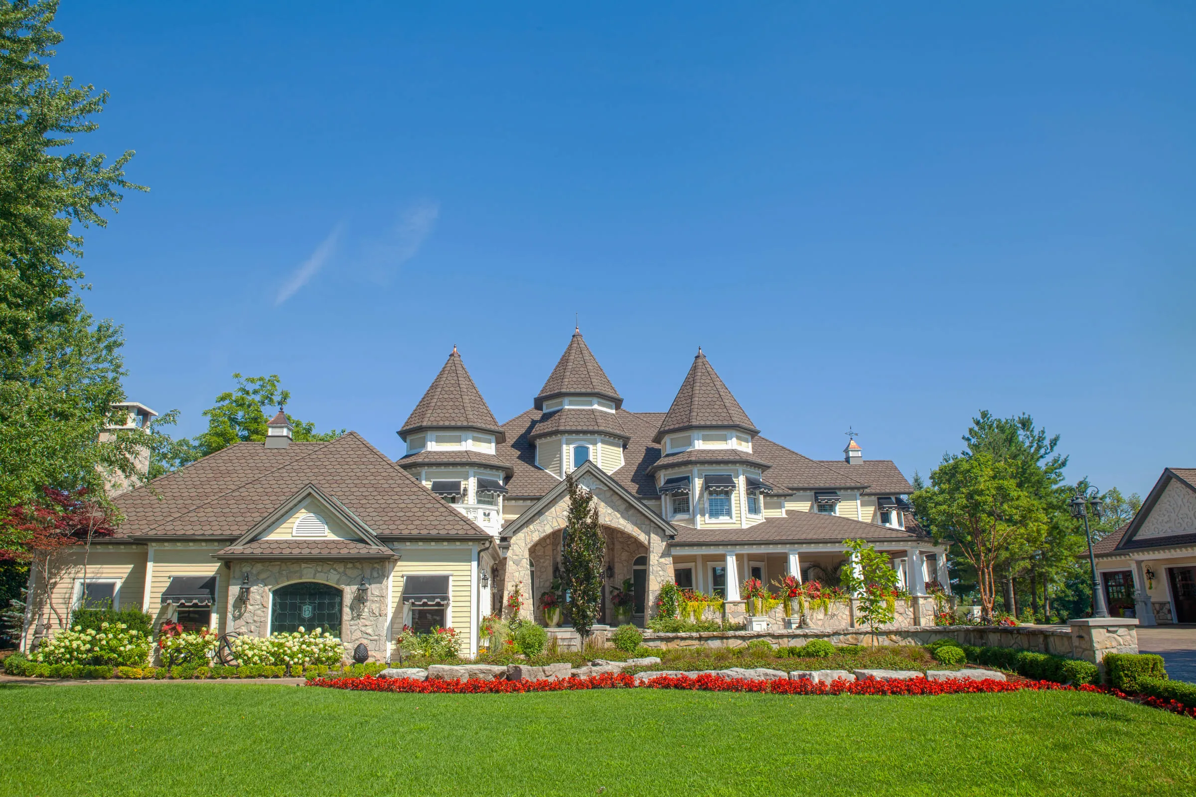 A large turreted house with manicured lawn and red flower beds under a clear blue sky