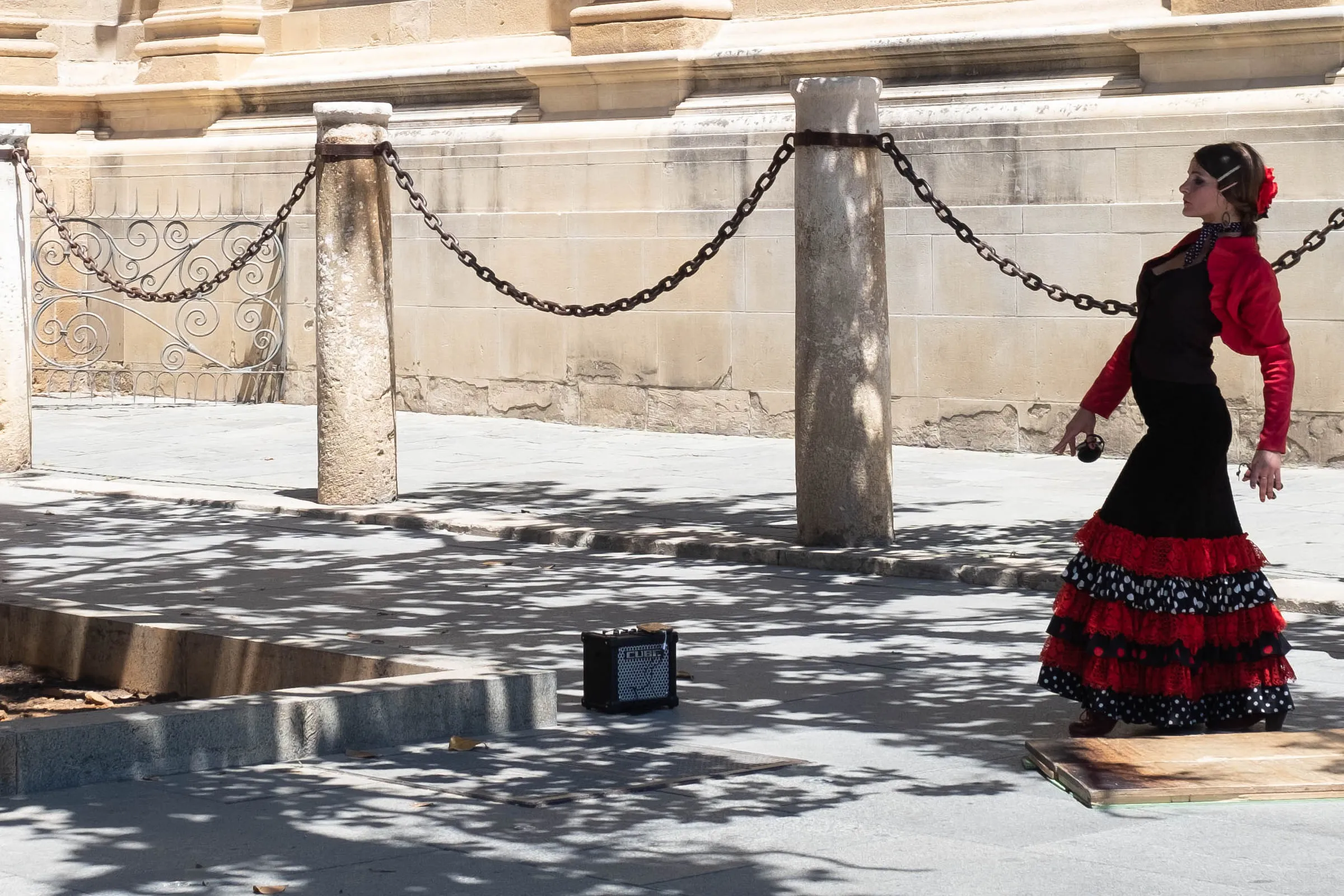 The same dancer in a black and red ruffled dress beside a chained stone bollard on a sunlit plaza
