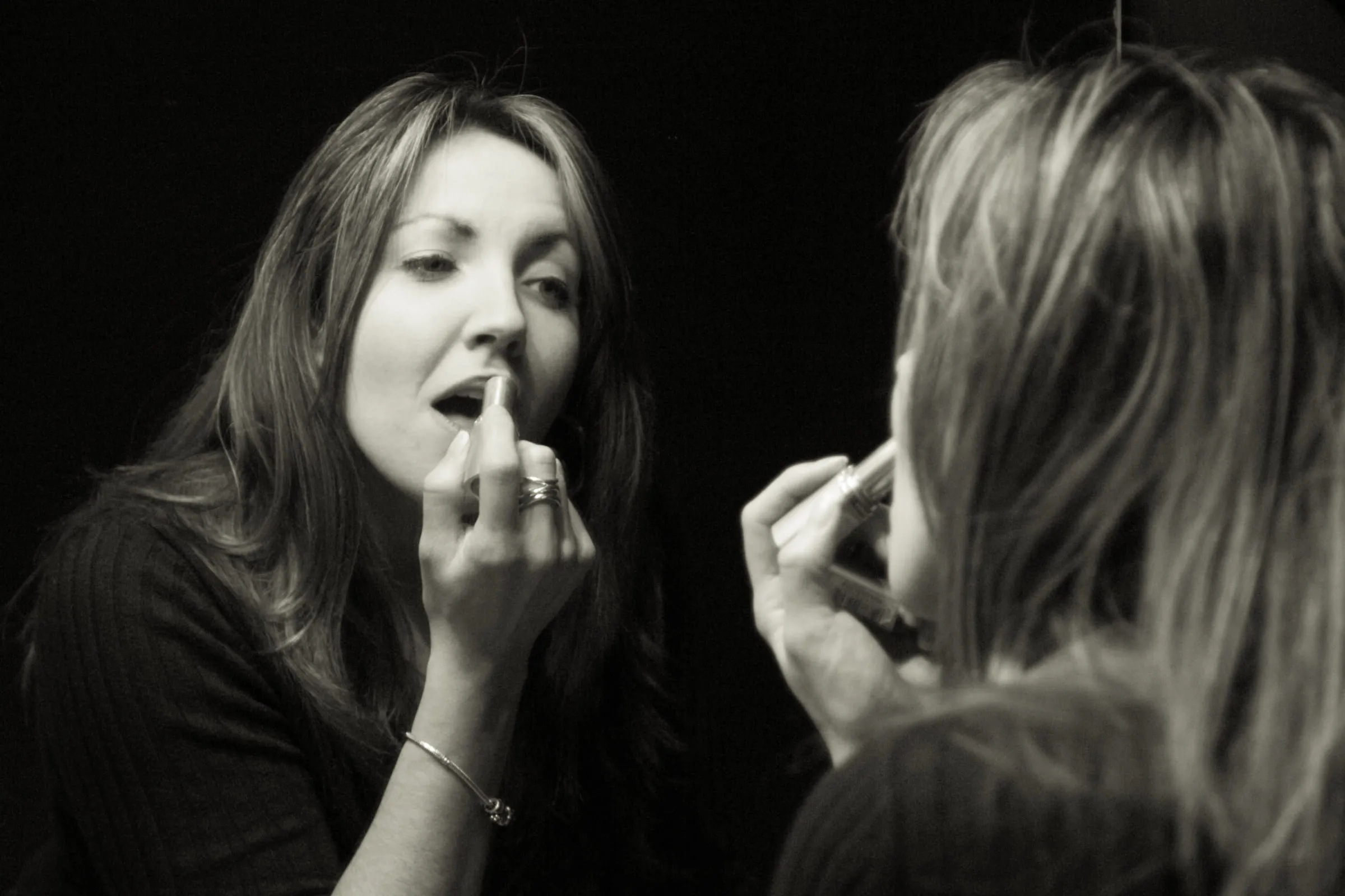 Black and white photograph of Mary applying lipstick at a mirror, her reflection alongside
