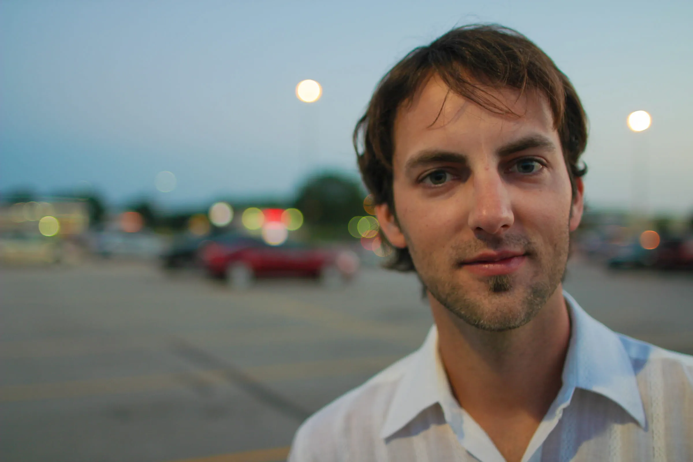 Portrait of Nathan in a parking lot at dusk, streetlights and car lights out of focus behind