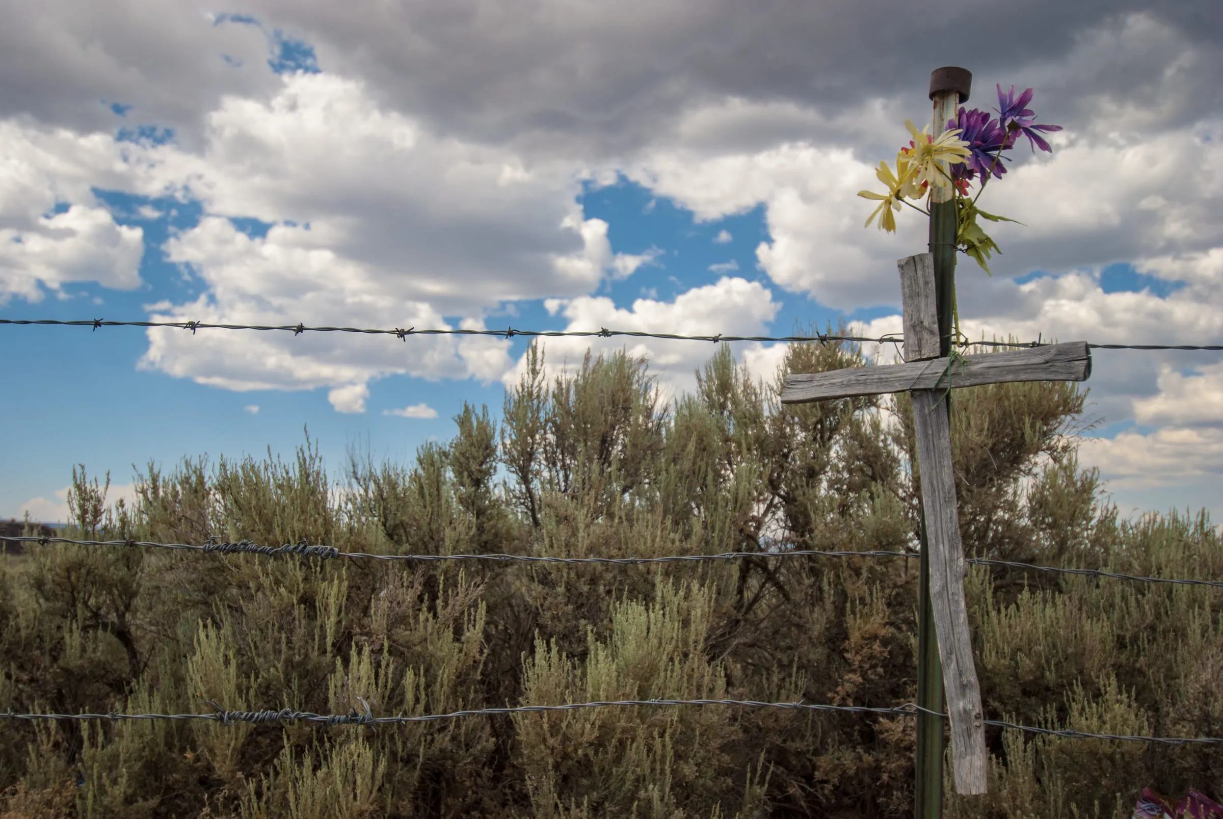 A weathered wooden cross topped with purple flowers behind a barbed wire fence in sagebrush