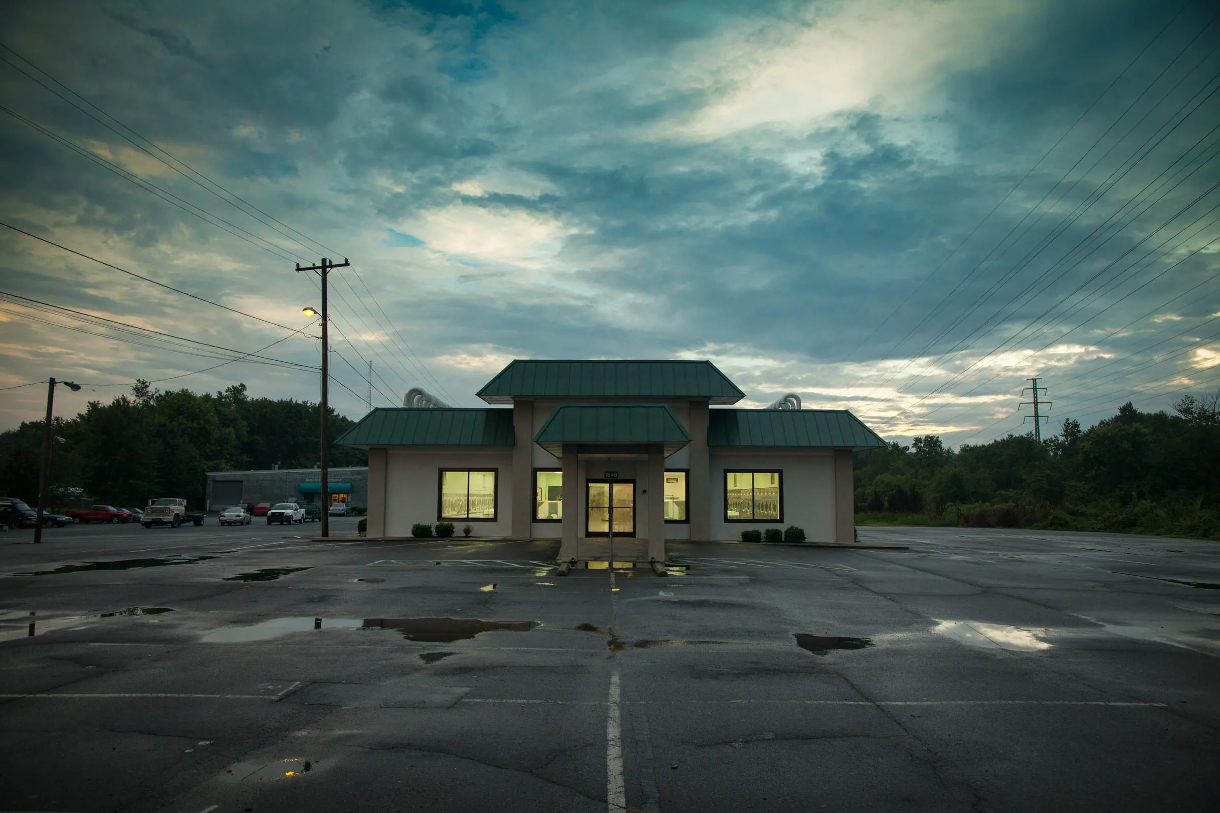 A lit laundromat at dusk across an empty rain-wet parking lot under a streaked grey sky