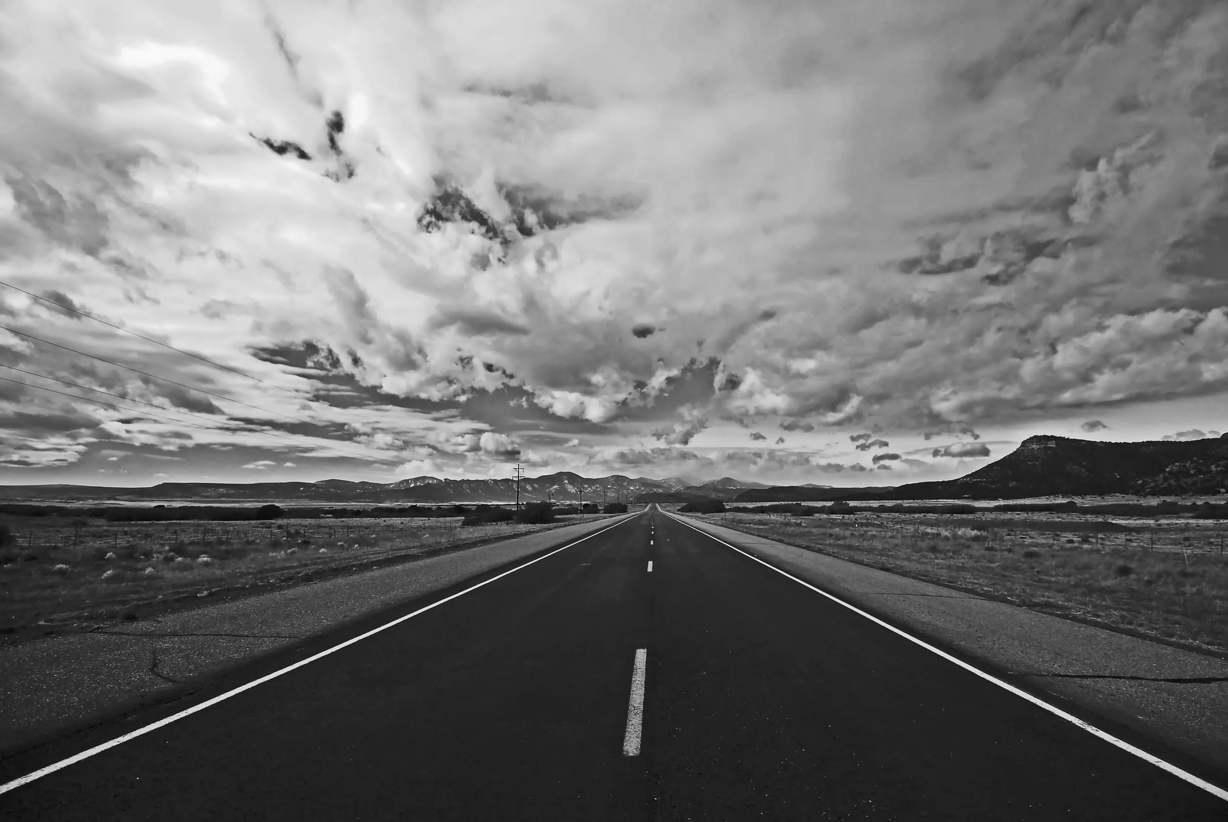 Black and white view straight down an empty two-lane highway toward distant hills under heavy cloud