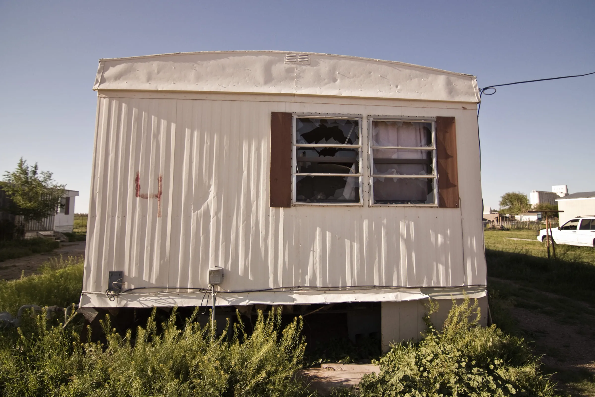 A white mobile home with a broken window and weeds grown up around its skirting