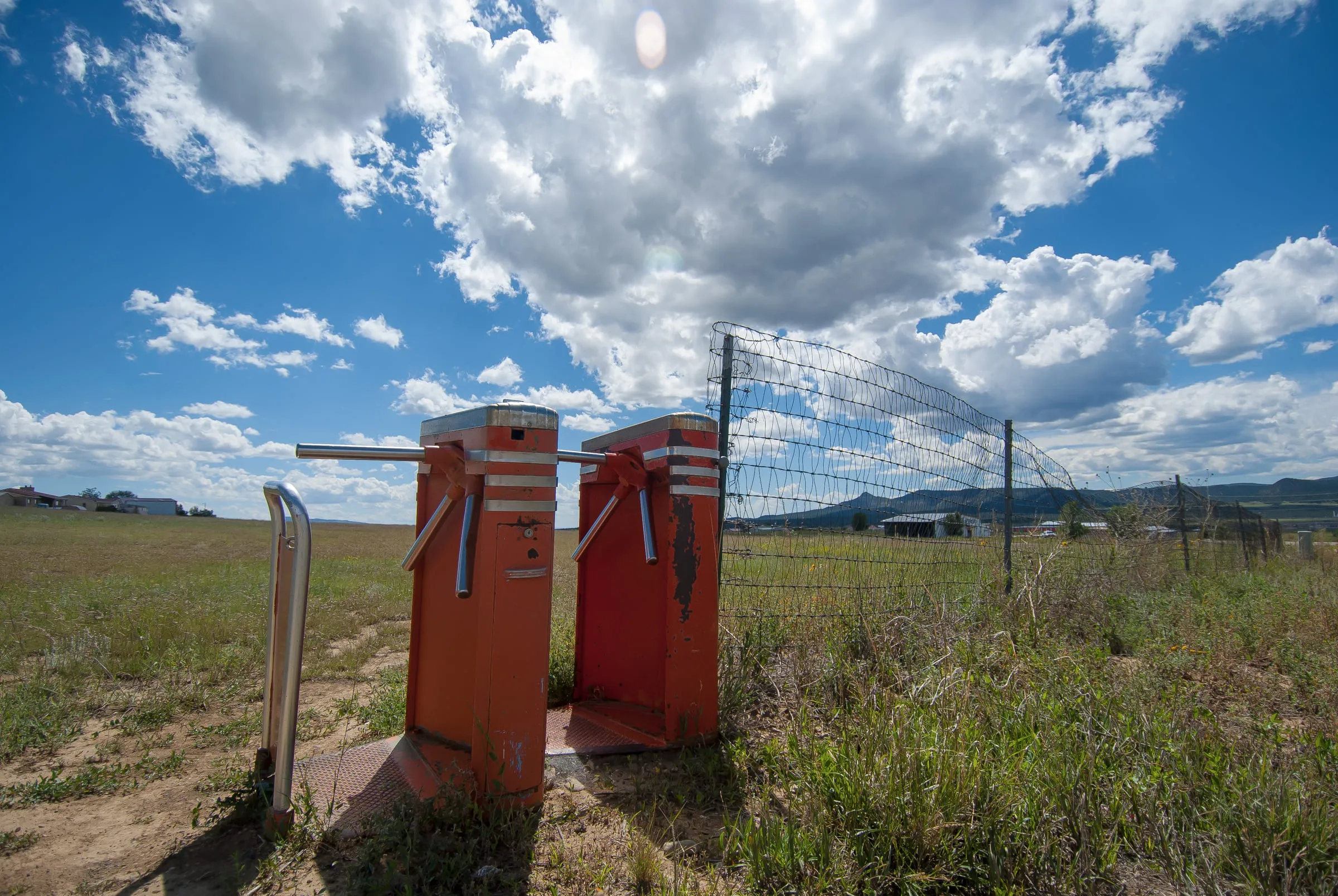Two red turnstiles standing alone in an overgrown field beside a chain-link fence